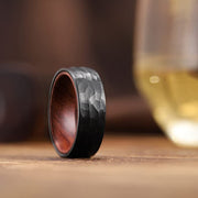 Hammered black tungsten ring with red wood interior on wooden table