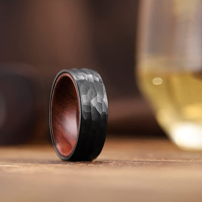 Hammered black tungsten ring with red wood interior on wooden table