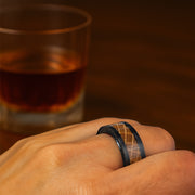 Hand wearing a black ring with a wooden inlay, glass of whiskey in the background